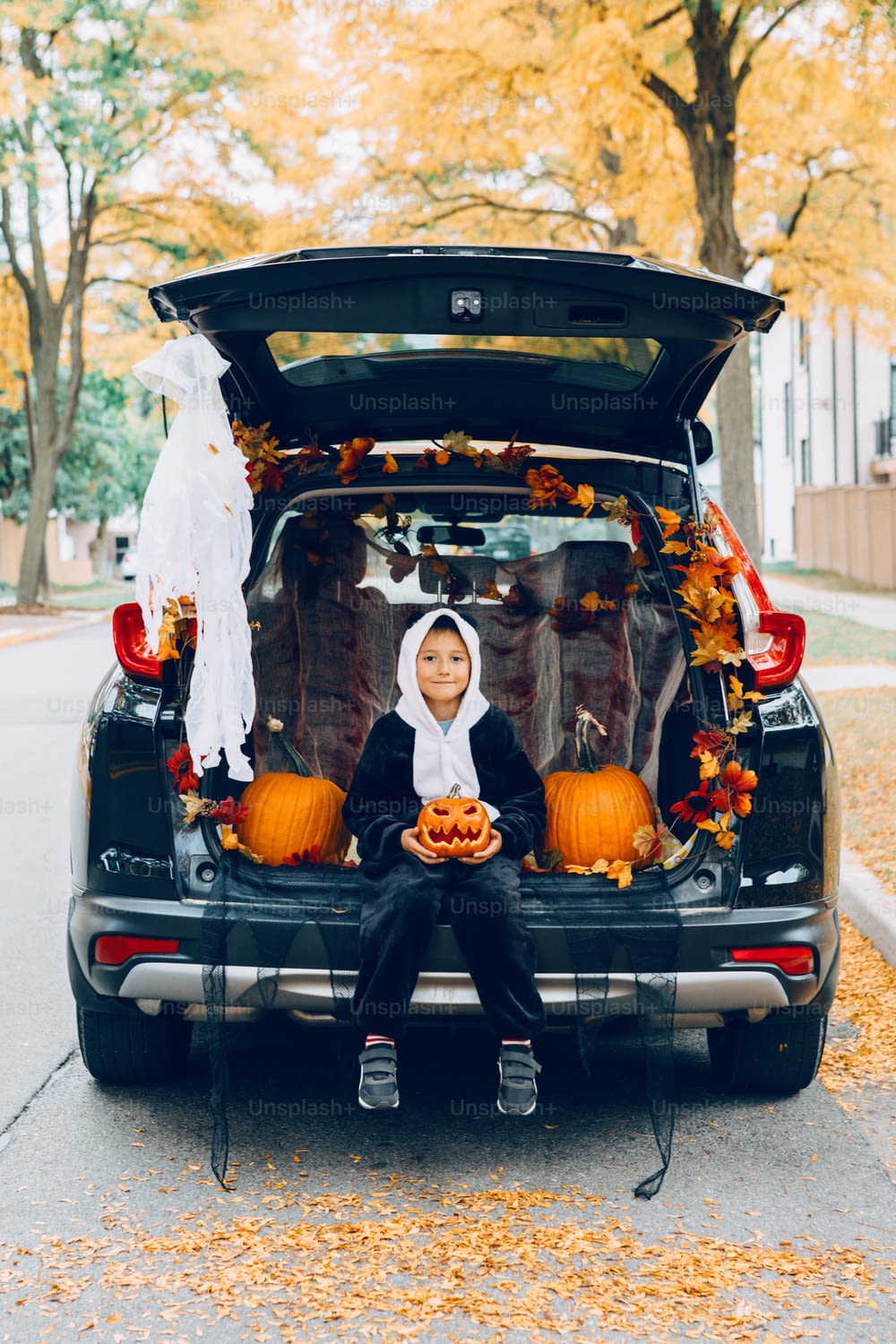 Truque ou tronco. Menino criança comemorando Halloween no porta-malas do carro. Criança com abóbora vermelha esculpida celebrando o tradicional feriado de outubro ao ar livre. Distanciamento social e celebração alternativa segura.