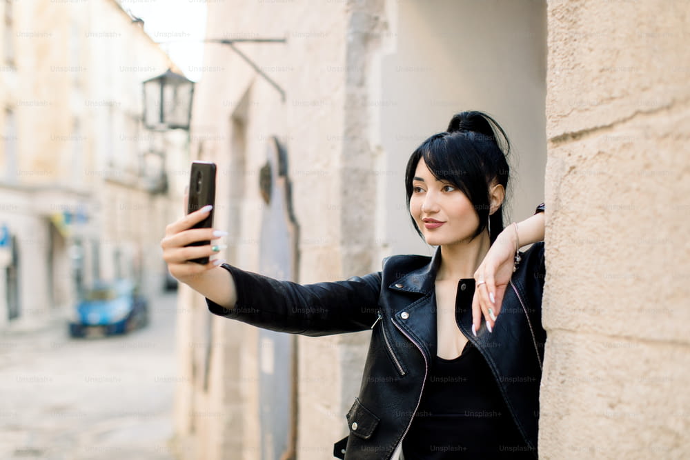 Foto urbana al aire libre de hermosa mujer sonriente asiática joven sexy con chaqueta de cuero negra, haciendo autorretrato en el teléfono inteligente, posando en la calle de la ciudad vieja. Selfie, gente, concepto de estilo de vida