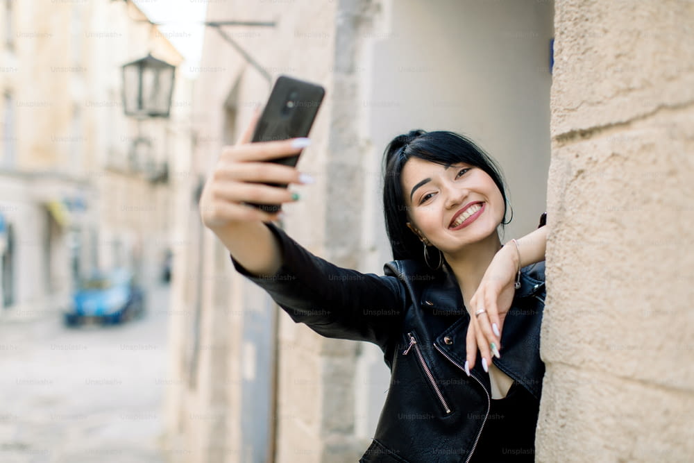Lifestyle portrait of young happy mixed raced Asian girl, using mobile phone, smiling, making selfie photo and having fun outdoors in the old city street. Morning city