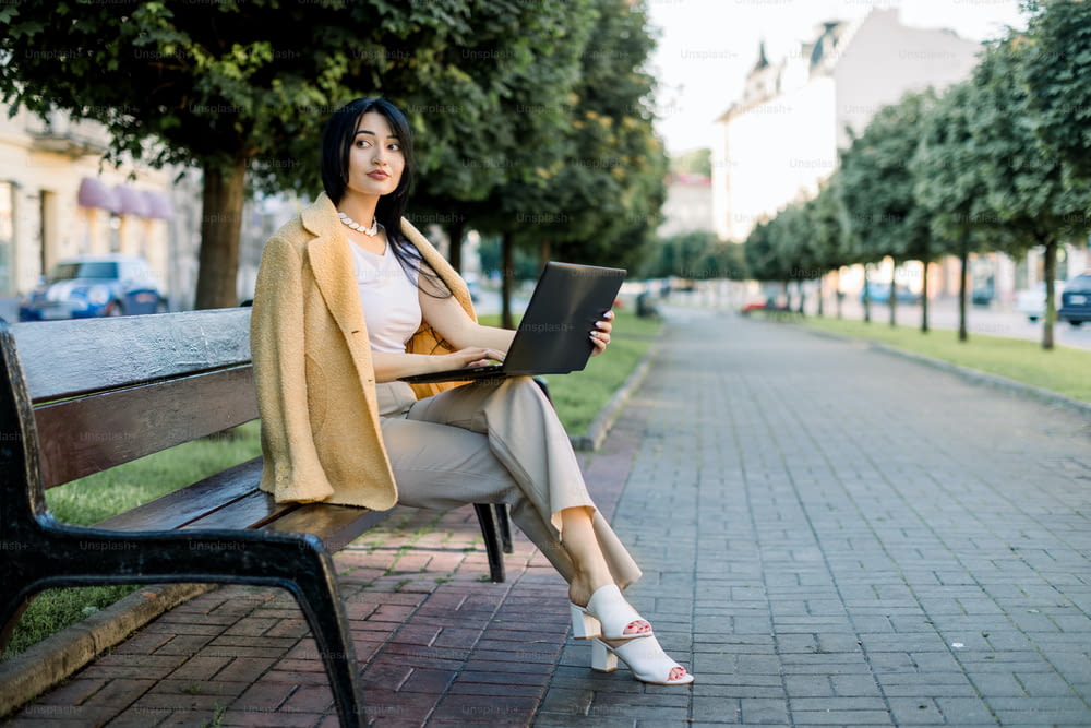 Junge hübsche, kluge, konzentrierte asiatische Geschäftsfrau, in elegantem, buntem gelbbeigem Anzug, sitzt mit ihrem Laptop auf der Stadtbank. Kostenloses WLAN in der Stadt, freiberufliches Arbeitskonzept.