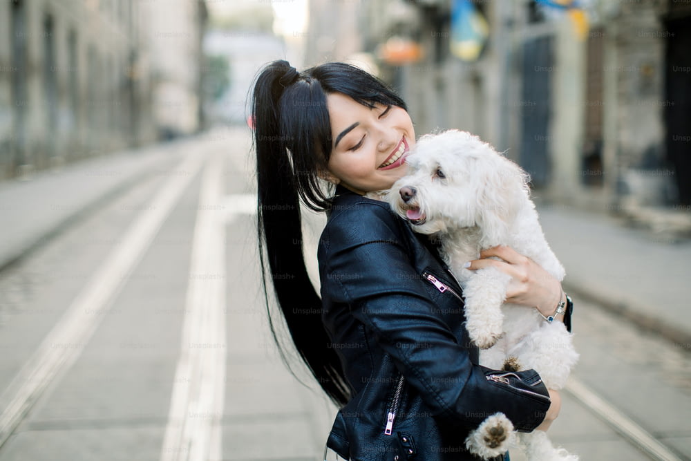 Una joven asiática de cabello oscuro, vestida con una chaqueta de cuero negro, se encuentra en la calle en la vieja calle de la ciudad europea, sosteniendo y abrazando al perrito blanco. Plano urbano horizontal, espacio de copia.