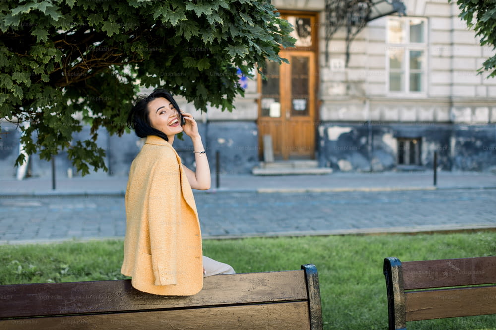 Laughing elegant Asian woman in yellow jacket, chilling and having fun during her morning city walk. Outdoor photo of lovely brunette business lady sitting on bench
