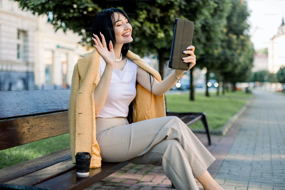 Happy young female Asian student or businesslady with a tablet and a disposable coffee cup, sitting on the bench in a summer city park, and having fun during video call, wavin to her friend.