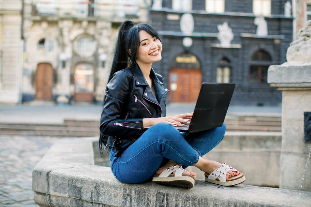 Wi-Fi gratuito en la ciudad, concepto freelance. Mujer asiática joven sonriente de raza mixta con cabello negro de cola de caballo sentada en la fuente y mensaje de texto en la computadora portátil, revisando noticias en las redes sociales