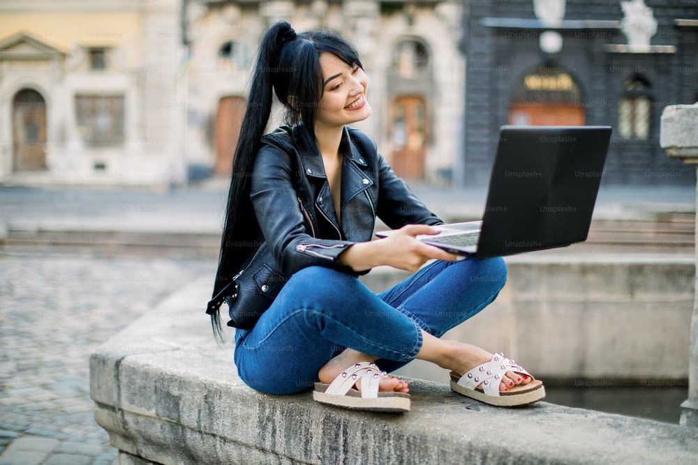 Heureuse hipster jeune femme métisse travaillant sur un ordinateur portable dans la ville. Belle femme asiatique aux cheveux en queue de cheval, assise sur une fontaine en pierre vintage, étudiant et utilisant un ordinateur portable