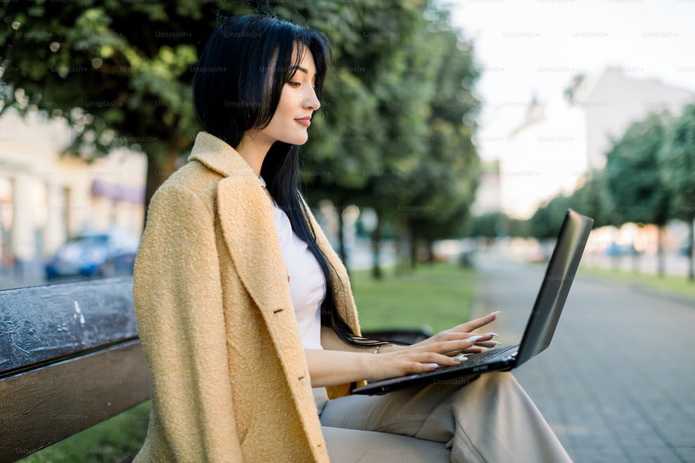 Retrato de una hermosa joven asiática, vestida con un colorido traje amarillo, usando una computadora portátil, leyendo noticias o sirviendo Internet, mientras está sentada en un banco en el callejón de la ciudad con árboles verdes.