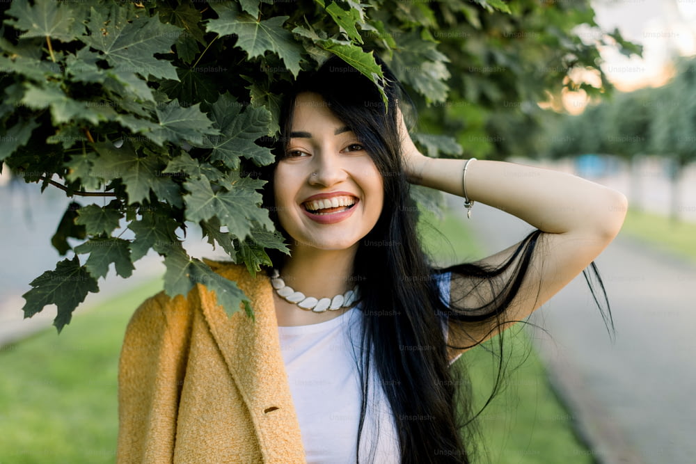 Jeune belle fille brune asiatique joyeuse portant un blazer jaune élégant et un t-shirt blanc, posant dans la rue de la ville sous l’érable, souriant à la caméra. Concept de mode de printemps et d’été.