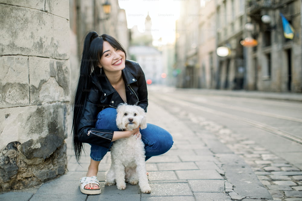 People, leisure, domestic animals concept. Beautiful young Asian woman, dressed in black leather jacket and jeans, walking with her dog in the city during the day. Copy space.