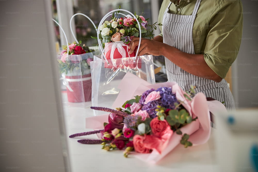 Cropped photo of a flower shop salesperson putting a pot of blooming flowers in a plastic bag near a bouquet