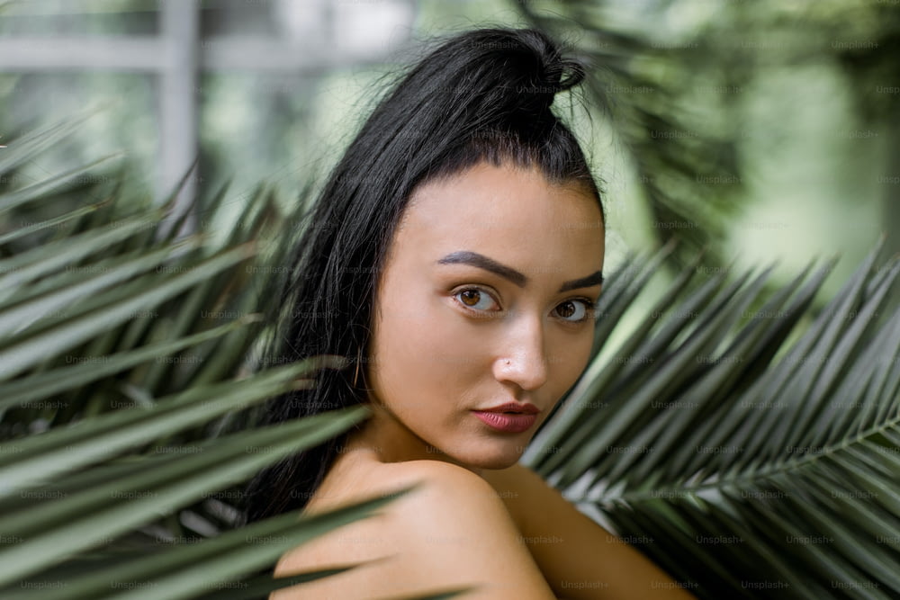 Close up beauty portrait of young charming Chinese Asian woman with ponytail hair style, posing to camera while standing near big exotic palm tree leaves in greenhouse or garden.