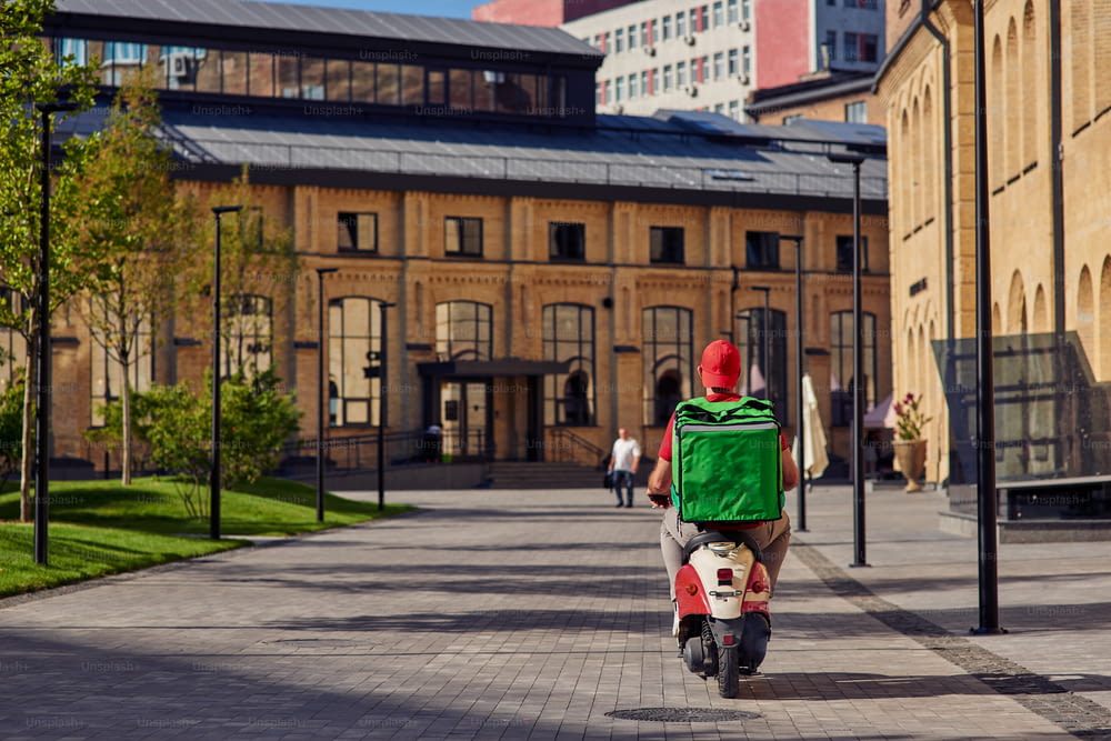 Rear view of courier with refrigerator bag riding scooter along beautiful sunny street with small houses. Food delivery concept