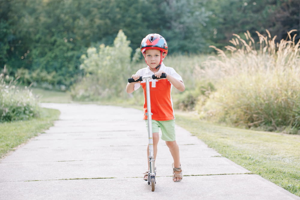 Glücklicher lächelnder kaukasischer Junge im Helm, der am Sommertag Roller auf der Straße im Park fährt. Saisonale Outdoor-Aktivitäten für Kinder. Gesunder Lebensstil in der Kindheit.