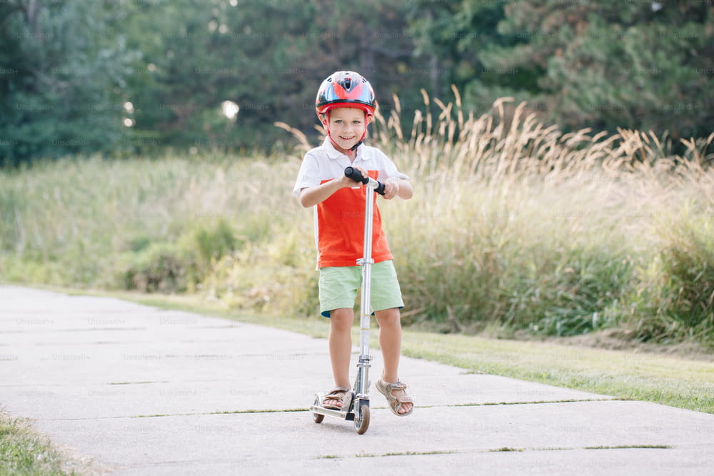 Felice ragazzo caucasico sorridente in casco che guida scooter sulla strada nel parco il giorno d'estate. Sport stagionale per bambini all'aperto. Stile di vita infantile sano.