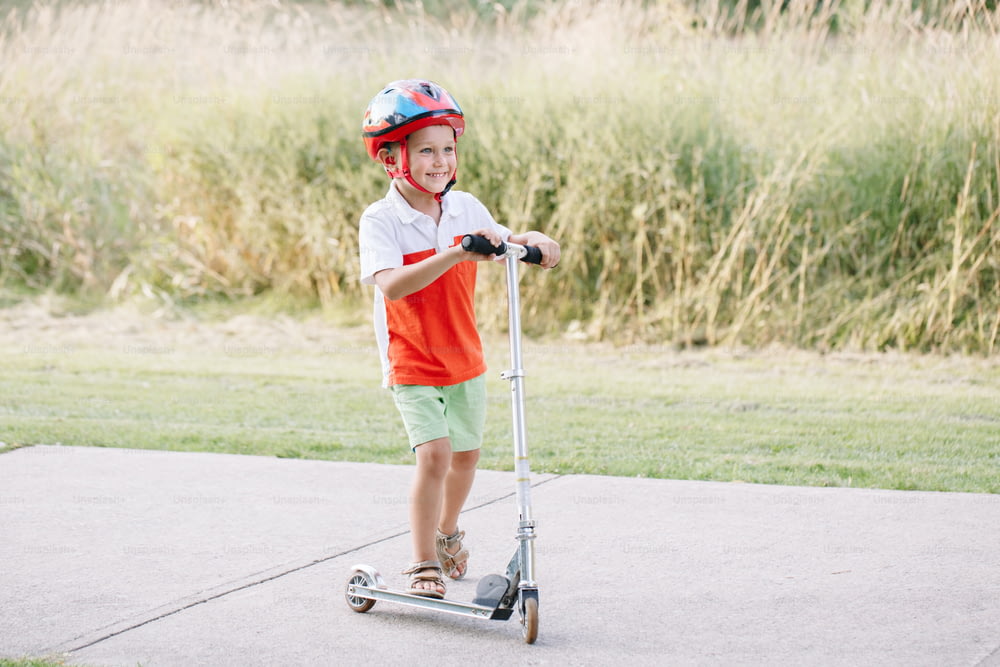 Felice ragazzo caucasico sorridente in casco che guida scooter sulla strada nel parco il giorno d'estate. Sport stagionale per bambini all'aperto. Stile di vita infantile sano.
