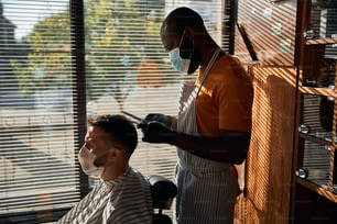 Afro American man barber wearing protective face mask and sterile gloves while cutting client hair with scissors