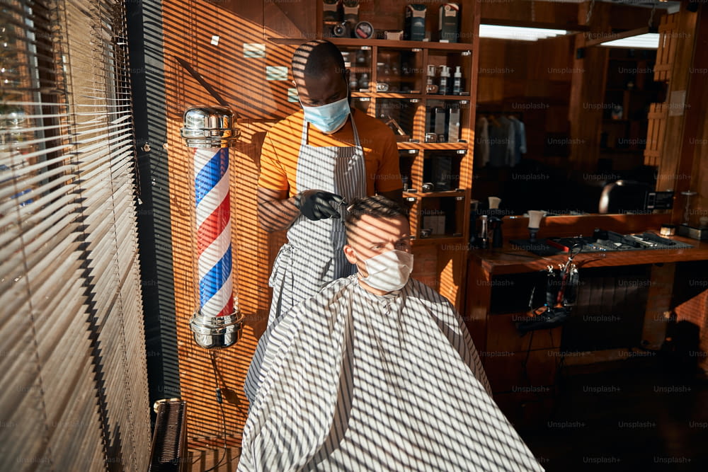 Handsome young man wearing protective face mask while getting haircut in barbershop during pandemic