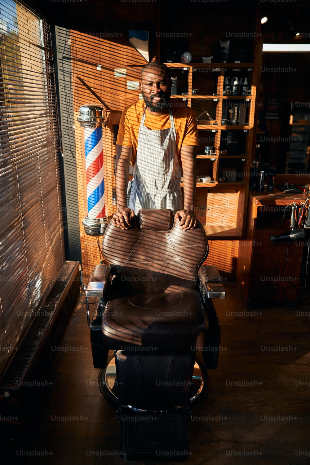 Handsome Afro American man in apron looking at camera and smiling while placing hands on barber chair