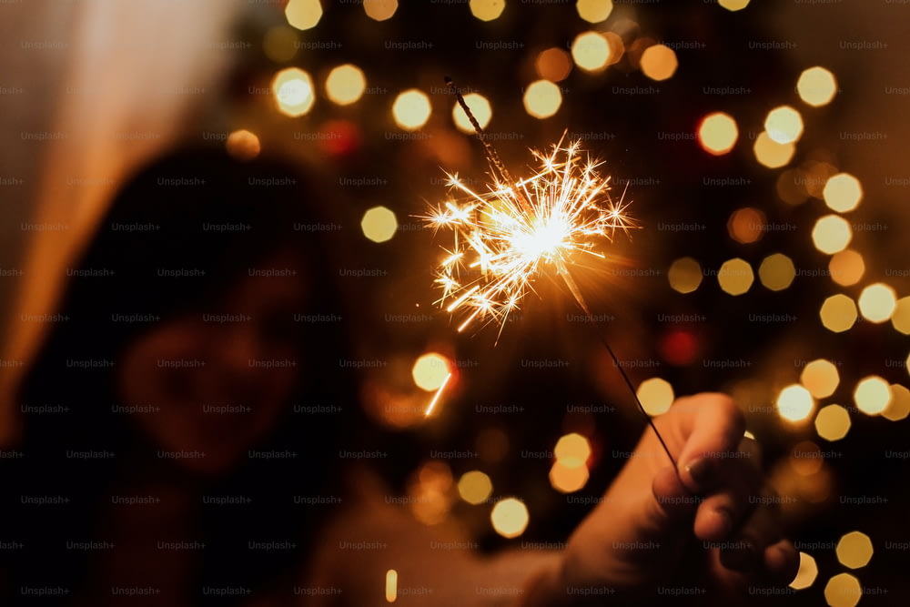 Young female celebrating new year with firework. Happy New Year! Stylish happy woman holding burning sparkler on background of  christmas tree with lights in dark festive room