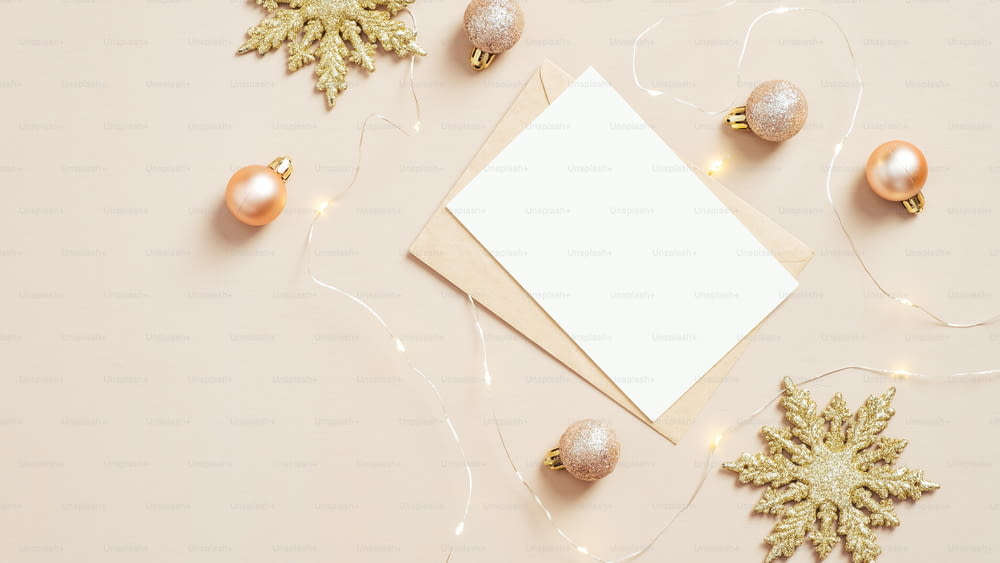 Paper envelope letter with blank white card mockup, golden snowflakes and balls on pastel beige background. Elegant Christmas flat lay composition, top view.