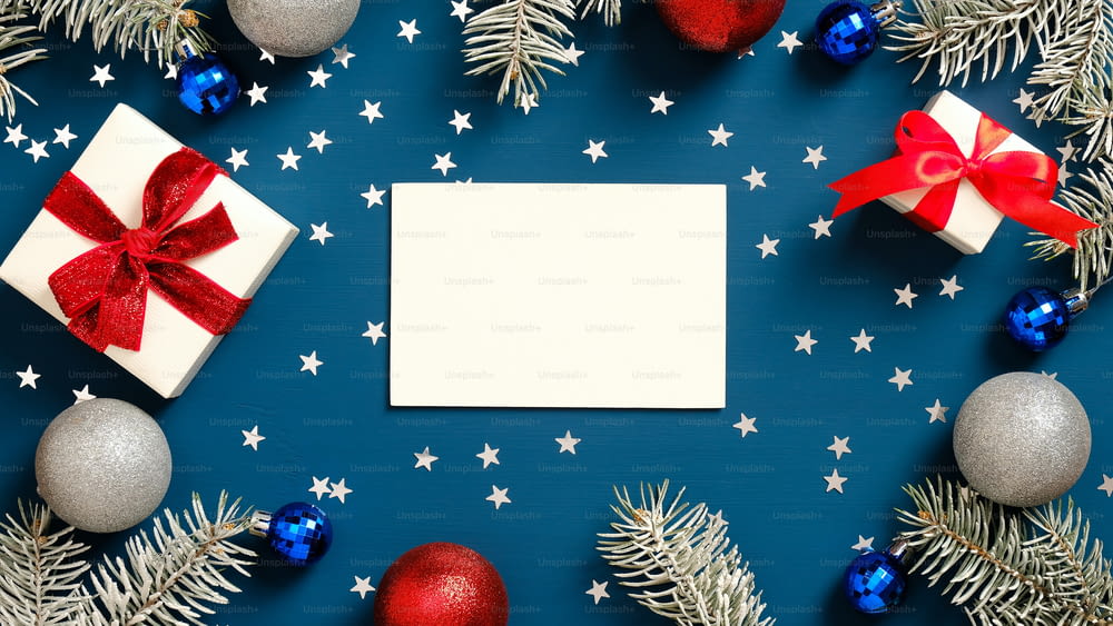 Merry Christmas greeting card mockup, white gift boxes with red ribbon bows, pine tree branches and decorations on dark blue background. Flat lay, top view.