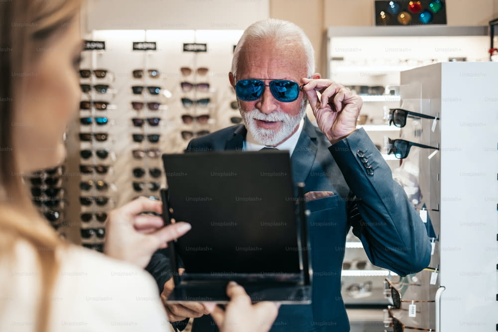 Élégant homme d’affaires senior choisissant et achetant des lunettes de soleil dans un magasin d’optique et jeune vendeuse l’aidant à prendre la bonne décision.
