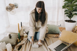 Femme élégante dans un pull confortable enveloppant une boîte-cadeau en tissu de lin dans une chambre bohème décorée de manière festive et regardant un ordinateur portable. Jeune femme regardant le tutoriel d’emballage cadeau furoshiki en ligne