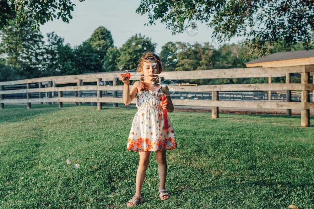Retrato sincero da menina caucasiana pré-escolar soprando bolhas de sabão no parque ao pôr do sol do verão. Autêntico momento de infância feliz. Estilo de vida crianças atividade sazonal ao ar livre.