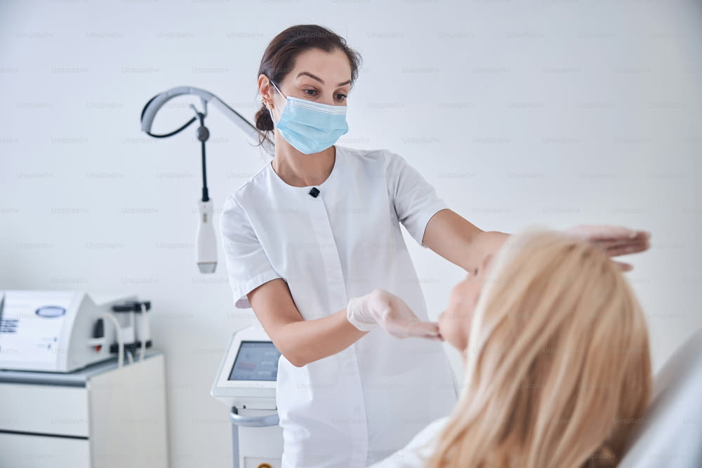 Side view portrait of professional beautician in white medicine uniform and mask looking to the skin face of elegant blonde woman in dermatological clinic