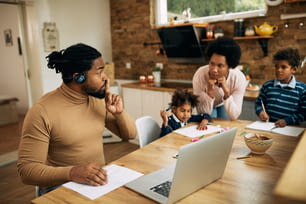 アフリカ系アメリカ人の男性が妻と子供を黙らせながら、ラップトップでビデオ通話をしたり、自宅で仕事をしたりしています。
