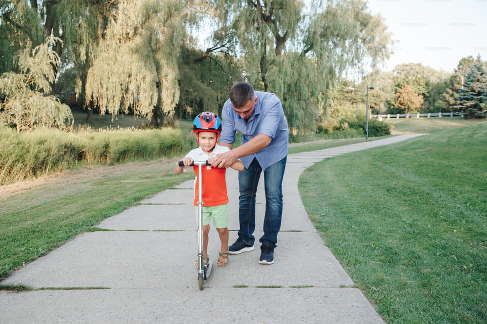 Pai caucasiano pai treinando ensinando filho menino a andar de patinete. Criança pré-escolar criança no capacete com bicicleta na estrada do parque do quintal. Atividade sazonal de verão para crianças ao ar livre.