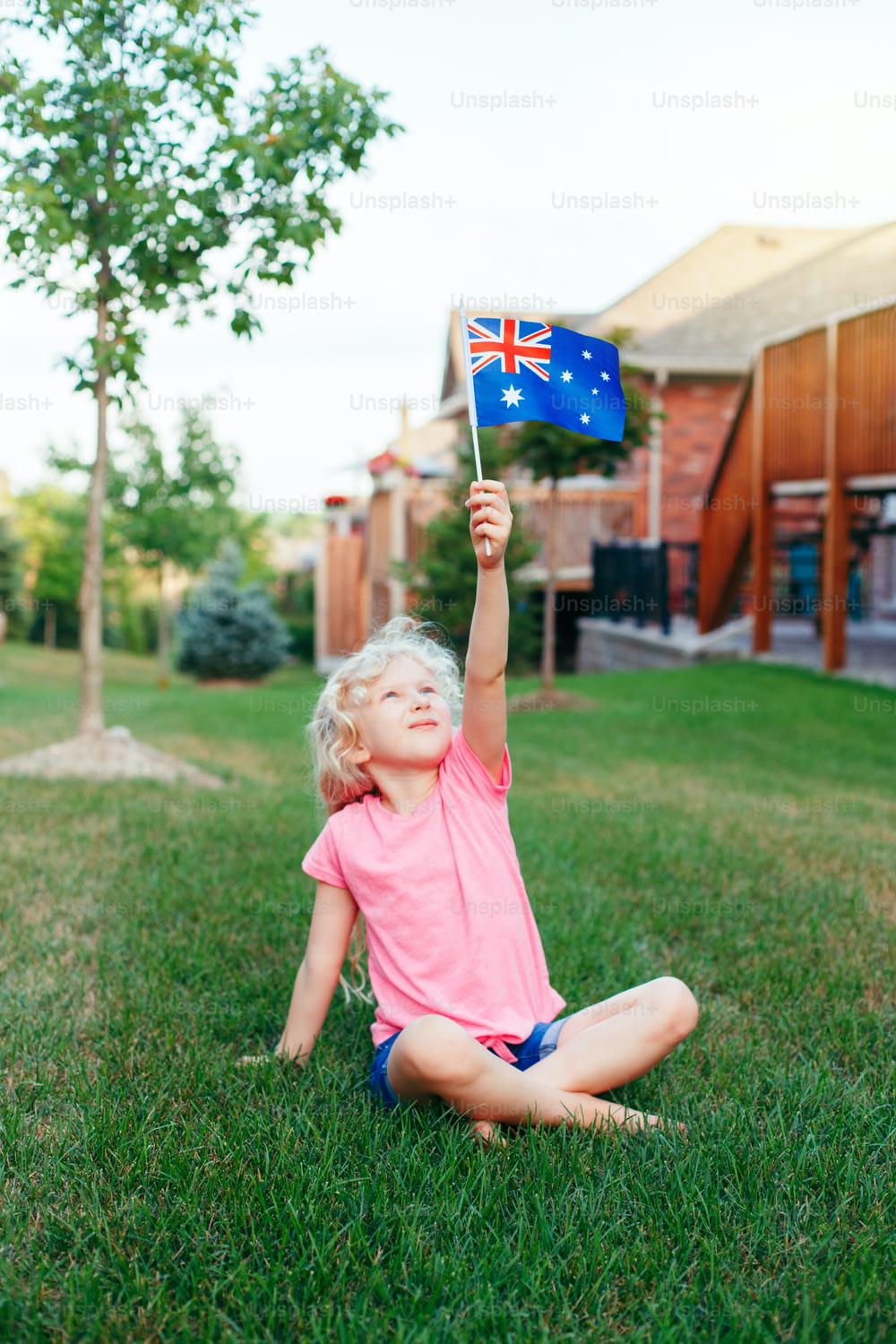 Happy Caucasian blonde girl holding Australian flag. Smiling child sitting on grass in park waving Australia flag. Kid citizen celebrating Australia Day holiday in January outdoor.