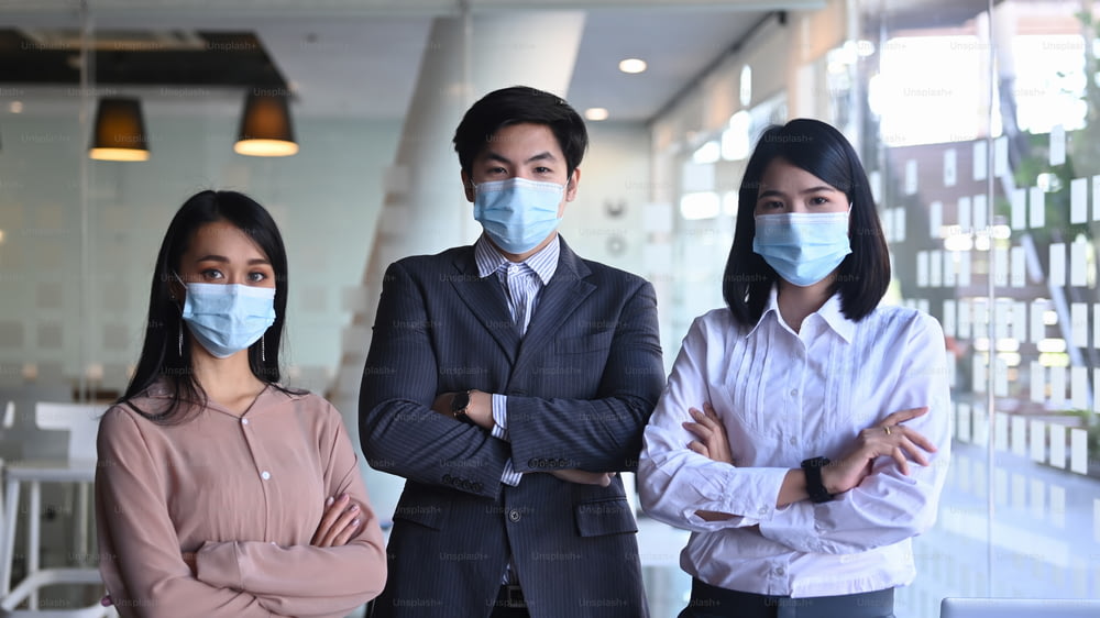 現代のオフィスで腕を組んで立っている間、医療用マスクを着用している自信のあるビジネスマン。
