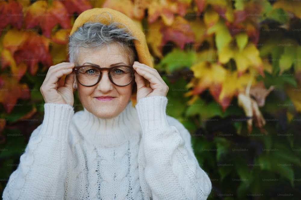 Portrait of senior woman standing outdoors against colorful natural autumn background, looking at camera.