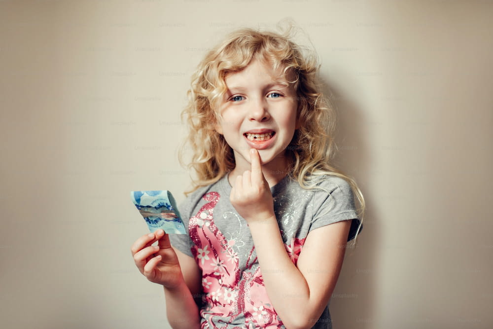Caucasian blonde girl showing her missing tooth in mouth and holding money from tooth fairy. Proud child kid showing lost tooth. Growing up stage and adolescence.