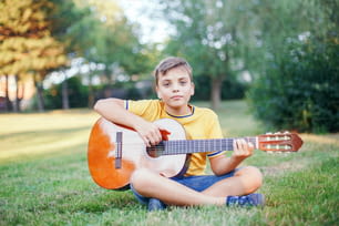 야외에서 기타를 연주하는 십대 초반의 소년을 듣기 어렵습니다. 귀에 보청기를 착용한 아이는 공원에서 음악을 연주하고 노래를 부릅니다. 어린이 어린이를위한 취미 예술 활동. 진정한 어린 시절의 순간.