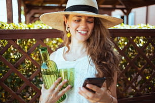sorridente elegante donna di 40 anni in camicia bianca con bottiglia di limonata, vetro e cappello che invia messaggi di testo utilizzando lo smartphone nel patio.