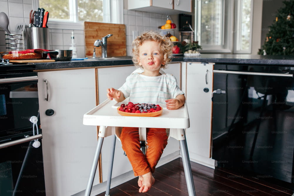 Lindo bebé caucásico comiendo frutos rojos maduros en casa. Divertido niño sentado en silla alta con bayas frescas en la cocina. Bocadillos saludables complementarios para niños pequeños.