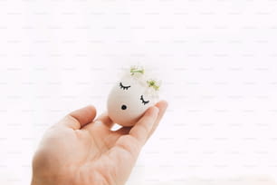 Hand holding easter egg with drawn sleeping face in floral wreath on white background in soft light. Happy Easter and Easter hunt concept ! Space for text. Eco friendly holiday