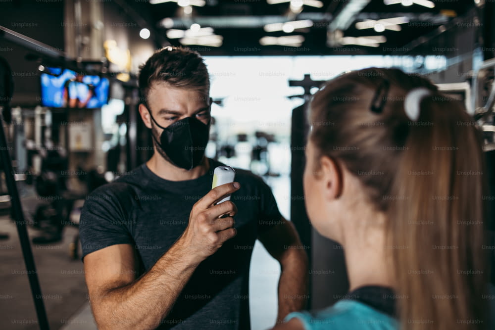 Young male fitness gym worker and instructor measuring body temperature of young sporty woman with infrared thermometer. Coronavirus world pandemic and sport theme.