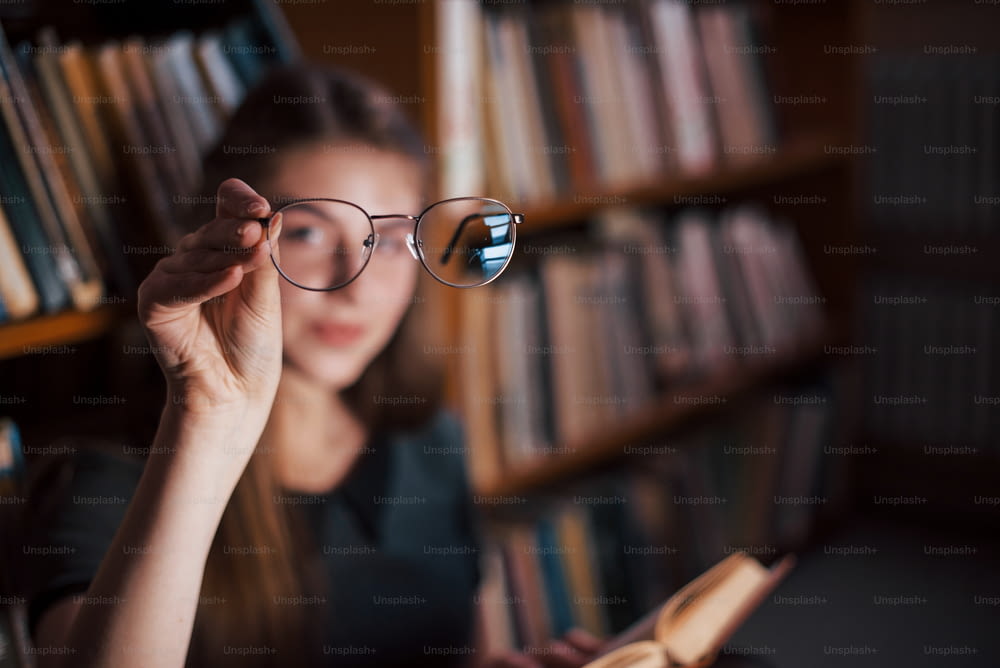 초점이 맞춰진 사진. 안경을 손에 들고 있습니다. 여학생은 책으로 가득 찬 도서관에 있습니다. 교육의 개념.
