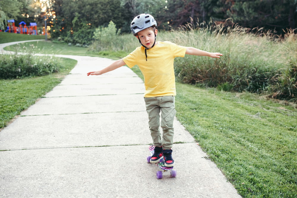 夏の日に公園の道路でスケートボードに乗っている灰色のヘルメットをかぶった幸せな白人の男の子。季節の屋外の子供活動スポーツ。健康的な子供時代のライフスタイル。スケートボードに乗ることを学ぶ男の子。