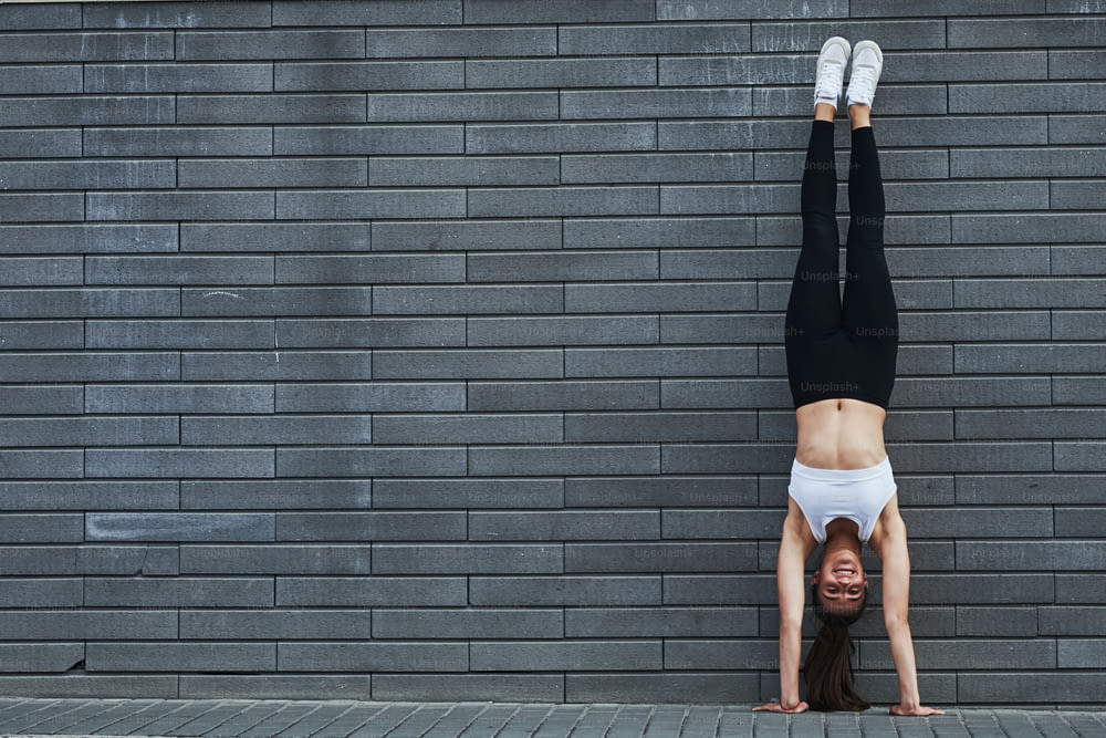 Doing handstand. Young sportive brunette with slim body shape against brick wall in the city at daytime.