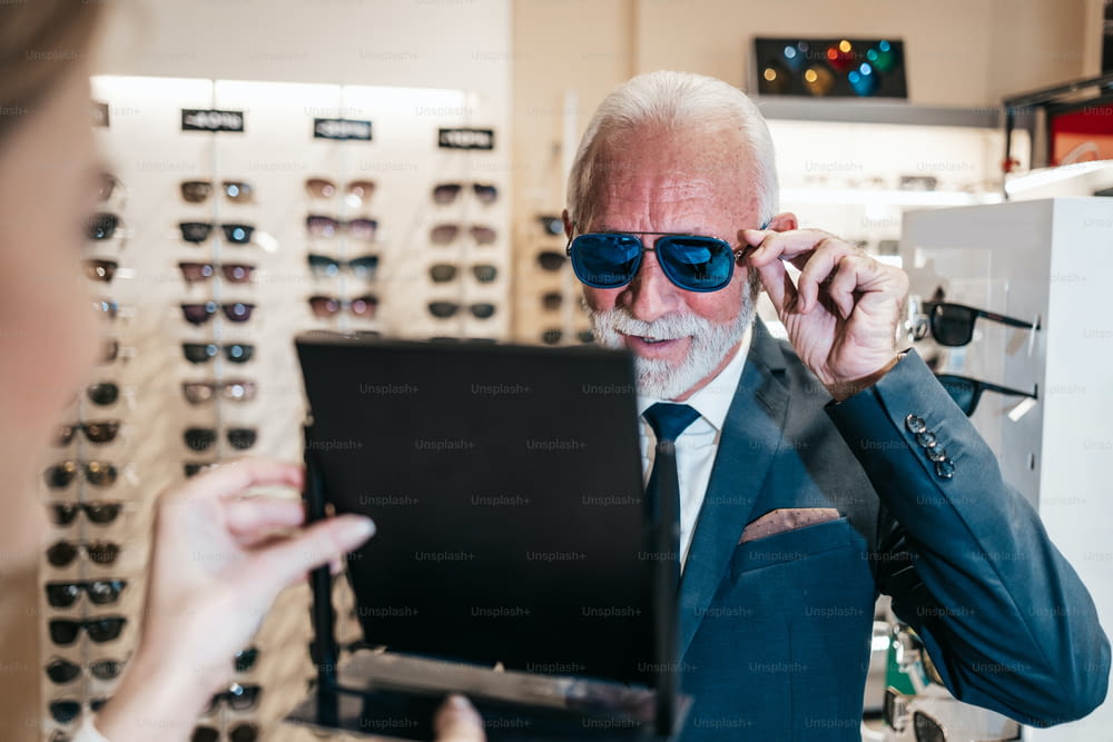 Élégant homme d’affaires senior choisissant et achetant des lunettes de soleil dans un magasin d’optique et jeune vendeuse l’aidant à prendre la bonne décision.