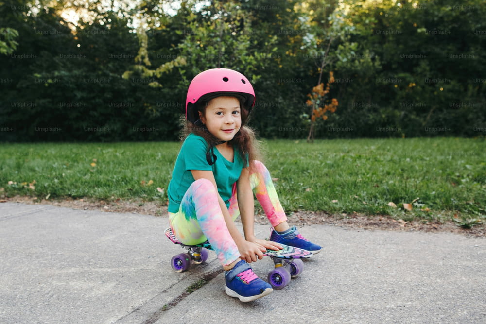 Happy smiling Caucasian girl in pink helmet sitting on skateboard on road in park at summer day. Seasonal outdoor children activity fun sport. Healthy childhood lifestyle.
