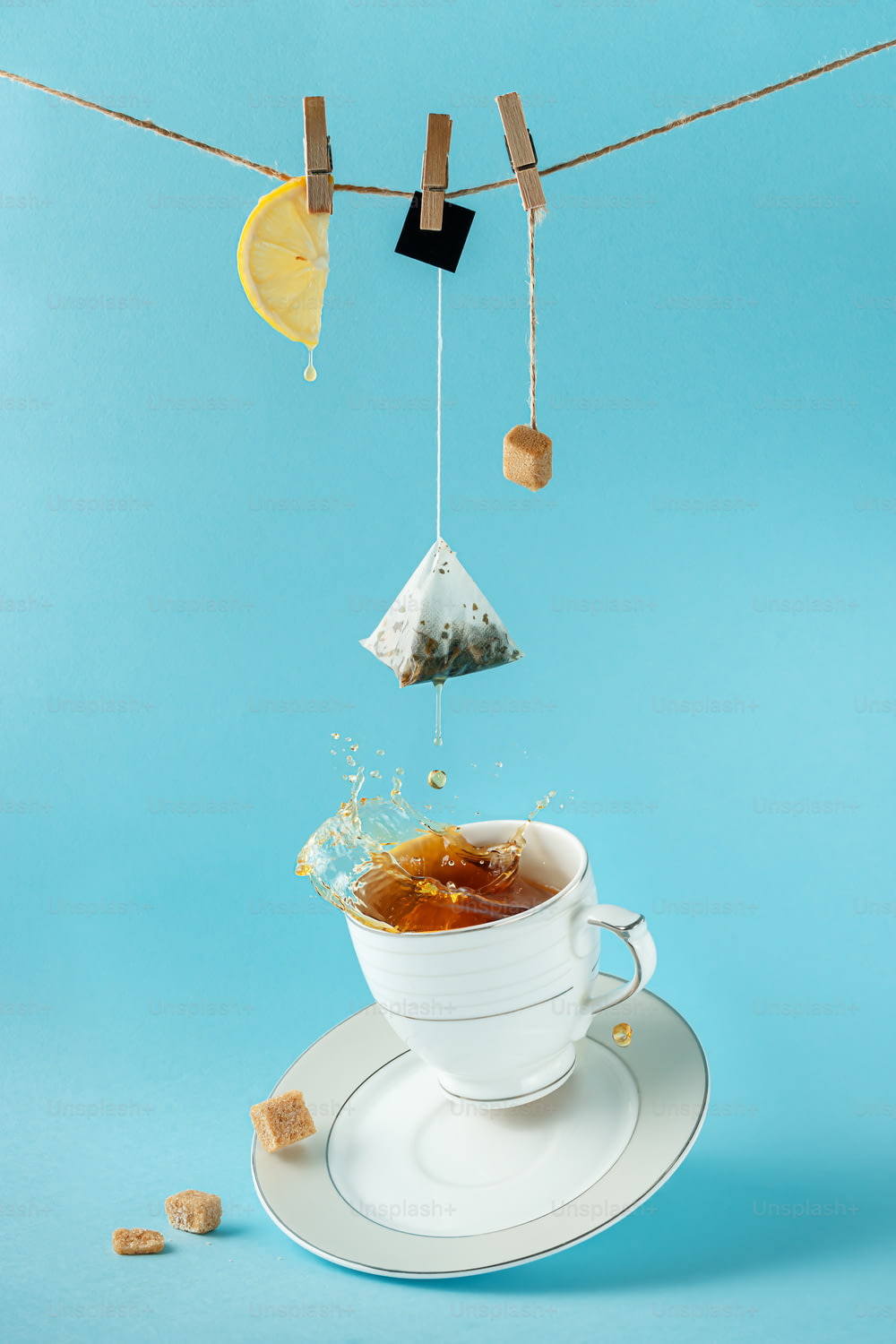 Saquinho de chá, limão e açúcar pendurados na corda sobre o chá espirrando na xícara em fundo azul. Natureza morta criativa