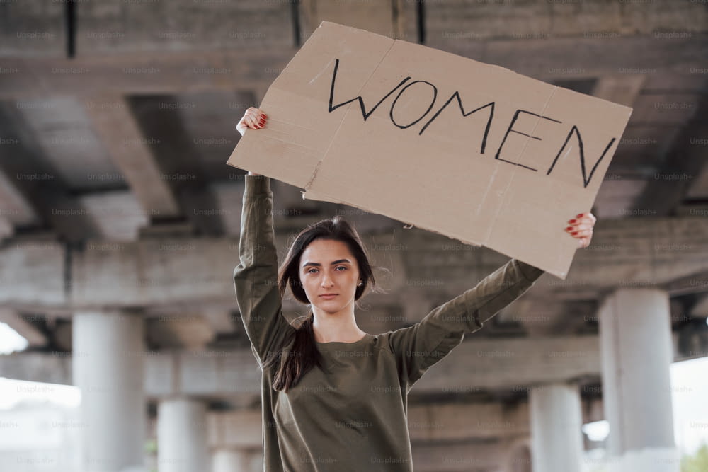 이 지역에서 혼자. 캐주얼 옷을 입은 예쁜 소녀가 손으로 만든 페미니스트 포스터를 손에 들고 서 있습니다.
