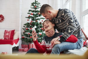 Un mec embrasse sa fille dans la tête alors qu’elle tient son smartphone. Des jeunes gens heureux s’assoient sur le rebord de la fenêtre dans la pièce avec des décorations de Noël.