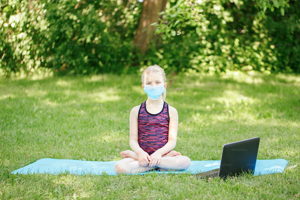 New normal. Yong girl child in face mask doing sport workout outdoors online. Video yoga class on Internet. Kid learning training in park with laptop. Social distance at coronavirus covid-19.