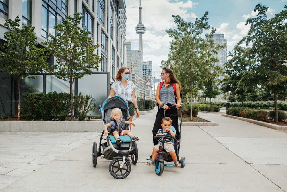 Two Caucasian moms with strollers and kids walking together in Toronto city Canada. Women in face masks with children outdoor. Friends talking on street keeping social distance. New normal.