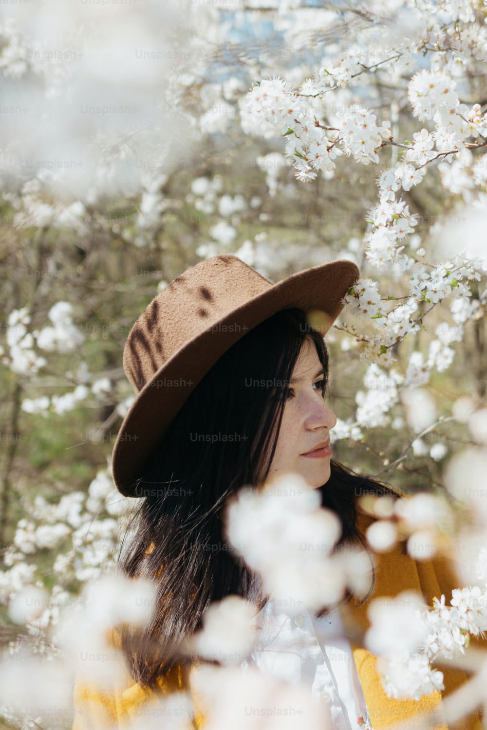 Stylish beautiful woman in hat sensually posing among blooming spring cherry branches outdoor,  calm moment. Portrait of fashionable young brunette female in yellow jacket embracing in white flowers